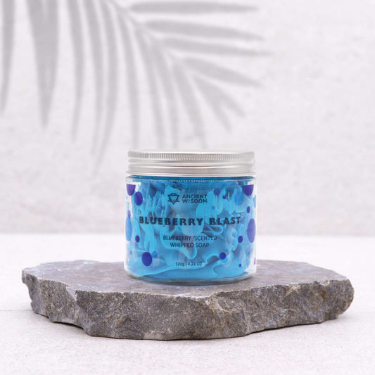 A jar of Blueberry Whipped Cream Soap placed on a rock, with a bright background and a shadow of a plant leaf cast upon it.